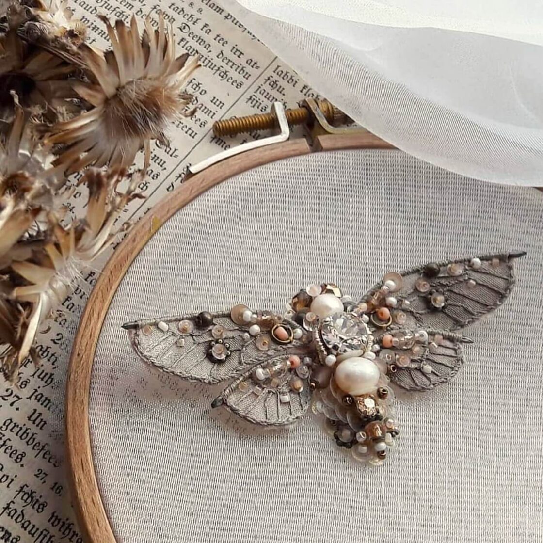 Hand-embroidered jewelry piece with insect motif and natural pearls on fabric, inspired by nature.